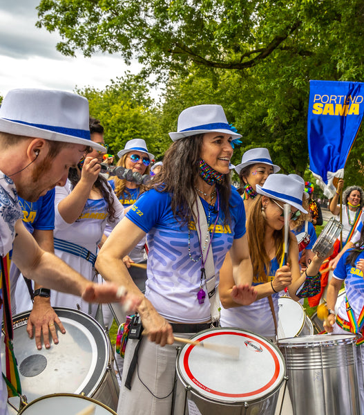 Group of Portland Samba players with blue and white t-shirts and white fedora hats. Crowd of people with drums and straps. Repique mor, repiques, surdos. Courtney Danley directing. Portland Samba banner in the background